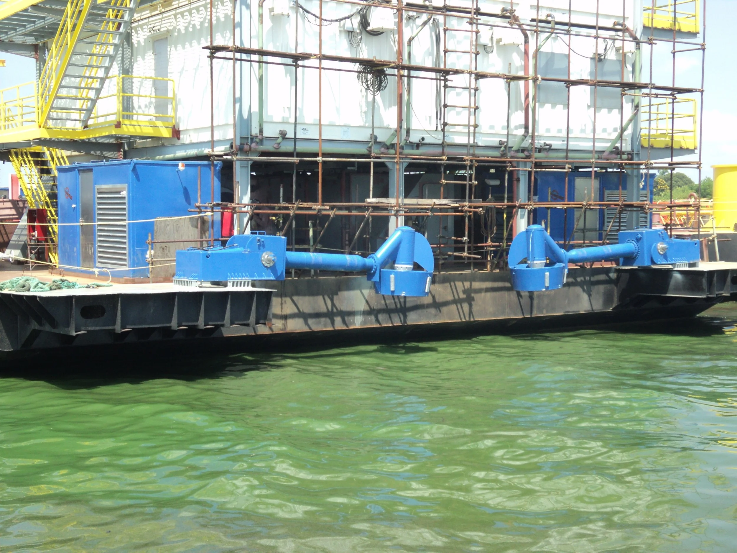 View of a barge with large blue pipes, surrounded by scaffolding, on green water at a construction or industrial site.