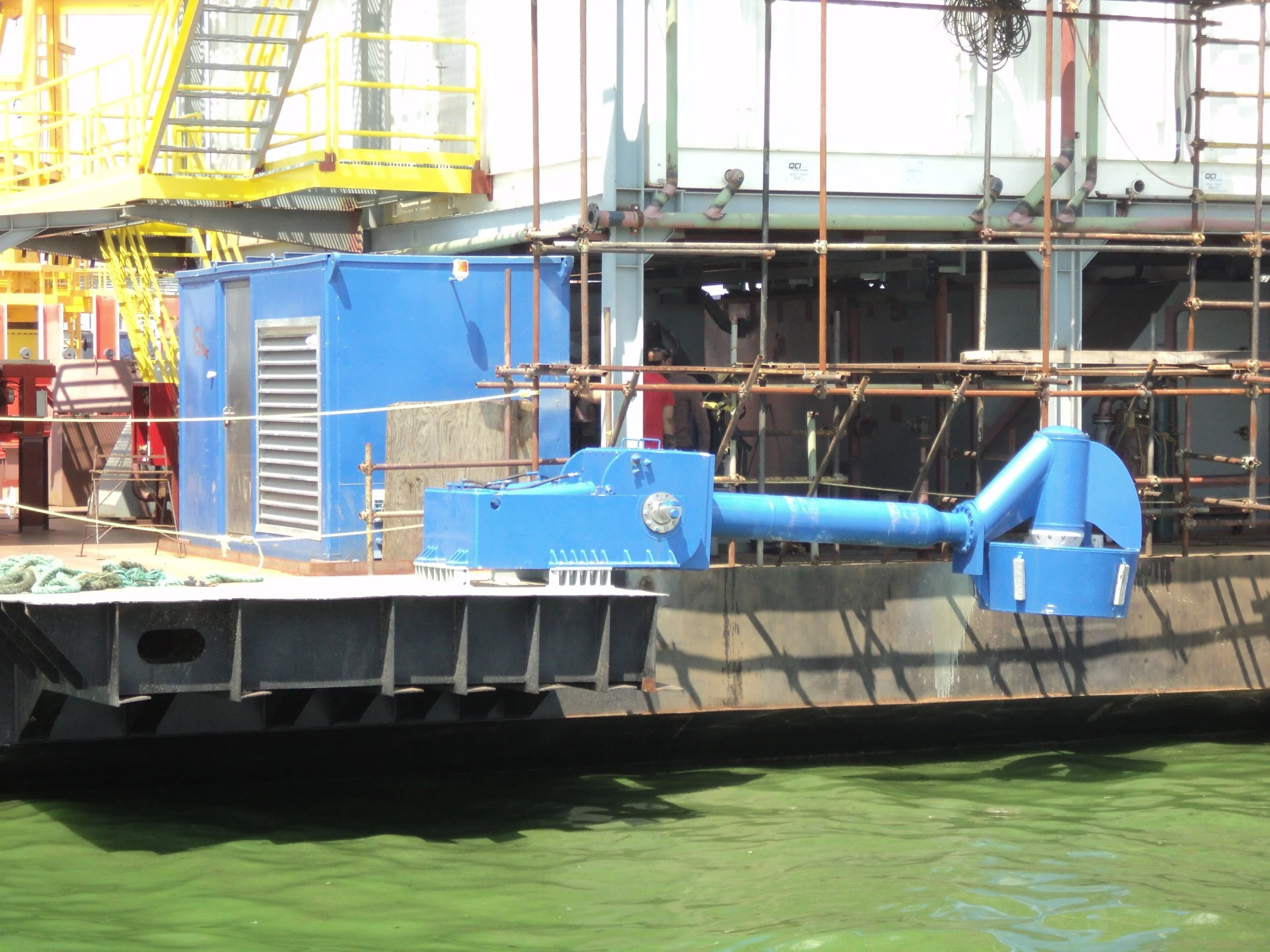 A ship's deck shown with a large blue exhaust vent and pipes, scaffolding, and industrial equipment, with green water in the foreground.