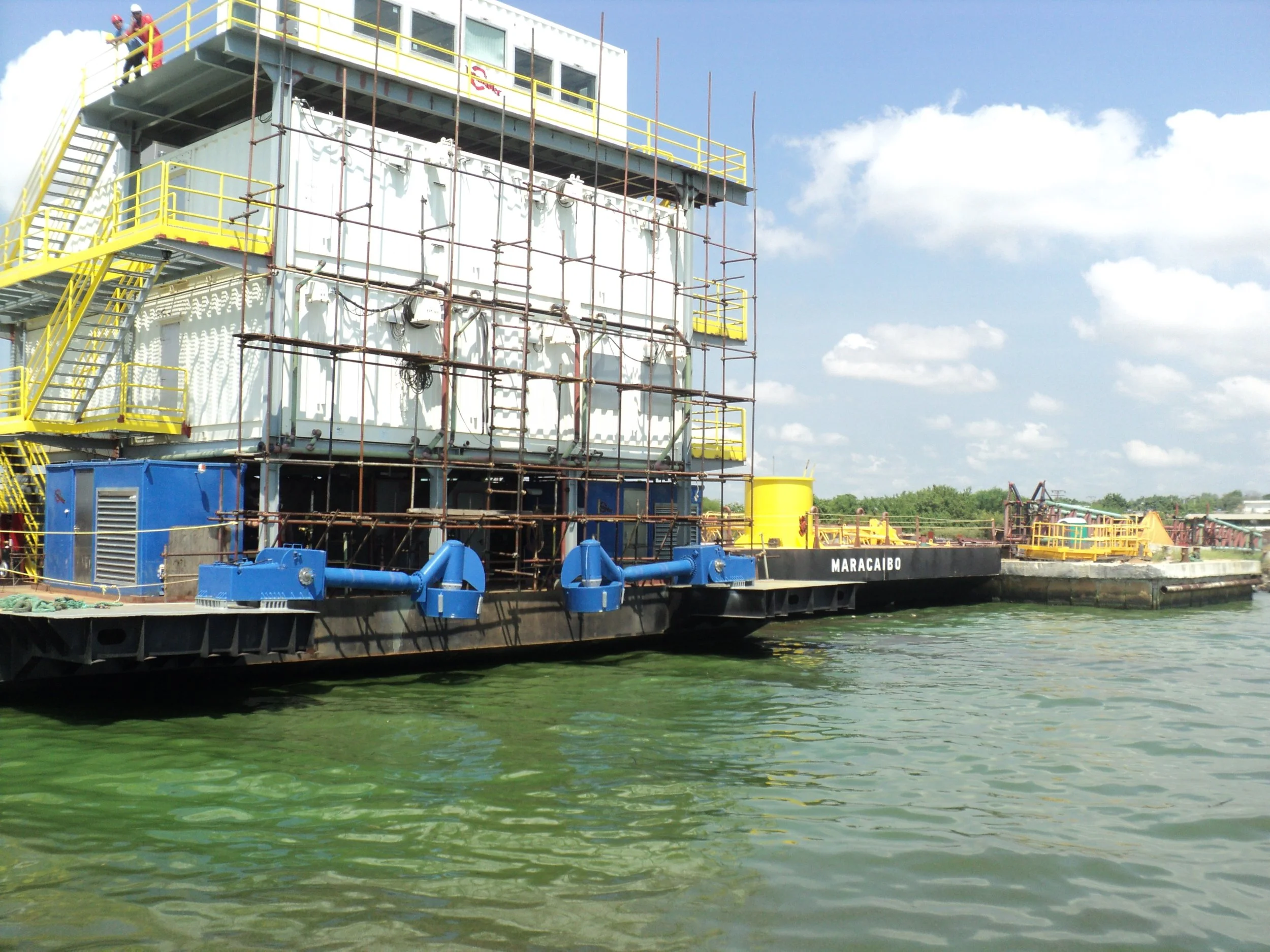 A large barge named Maracaibo on a body of water, with a multi-story structure underneath construction or maintenance, surrounded by scaffolding and yellow safety railings, under a partly cloudy sky.