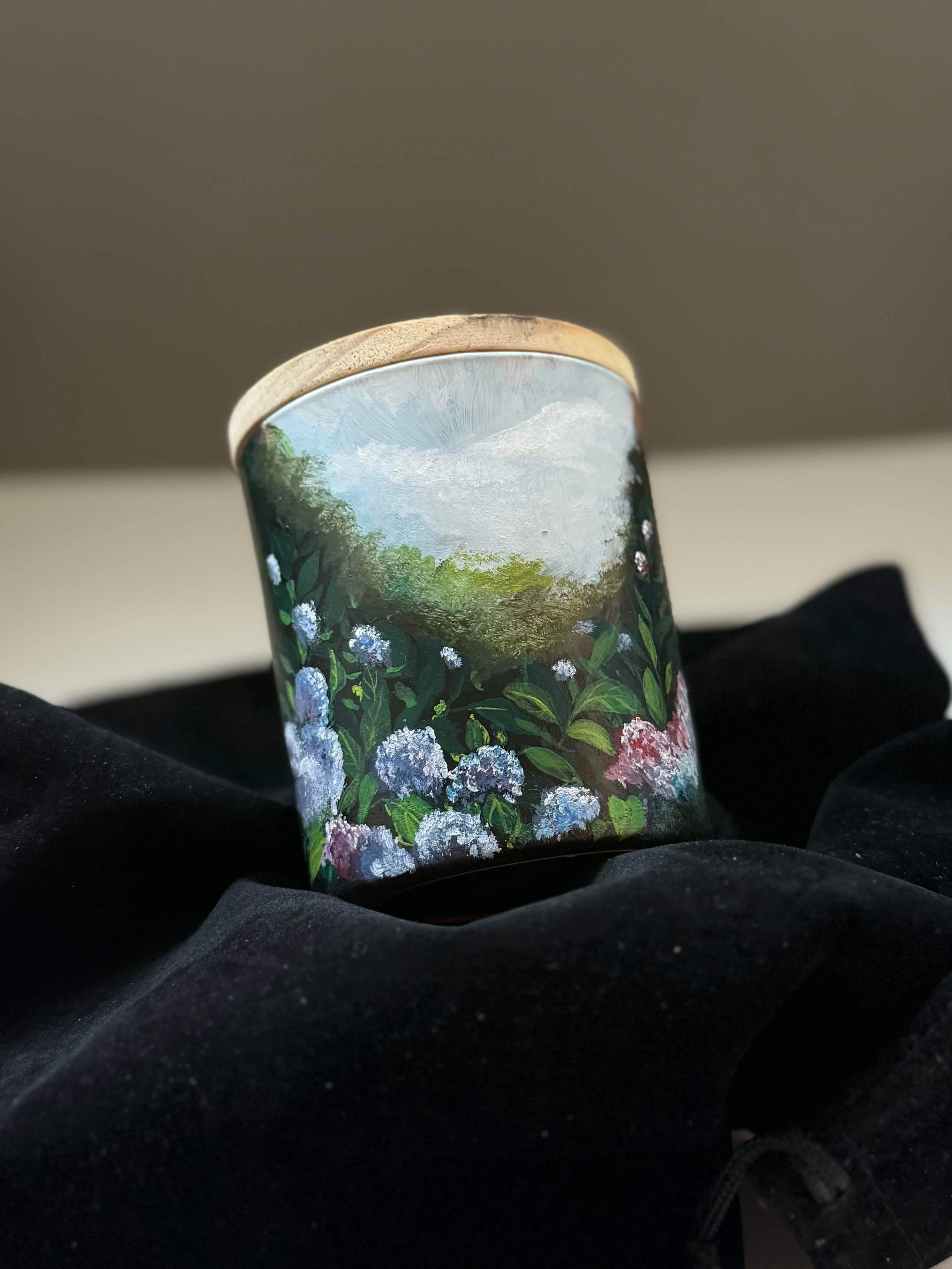 A hand-painted cylindrical box featuring a landscape scene with trees and blooming flowers, set against a cloudy sky background.