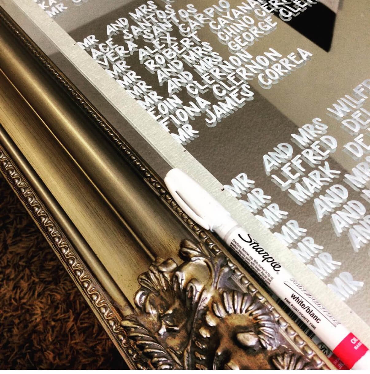 A decorative framed piece of art with white text on a black background, featuring names and titles, and a white dry erase marker resting on the frame.