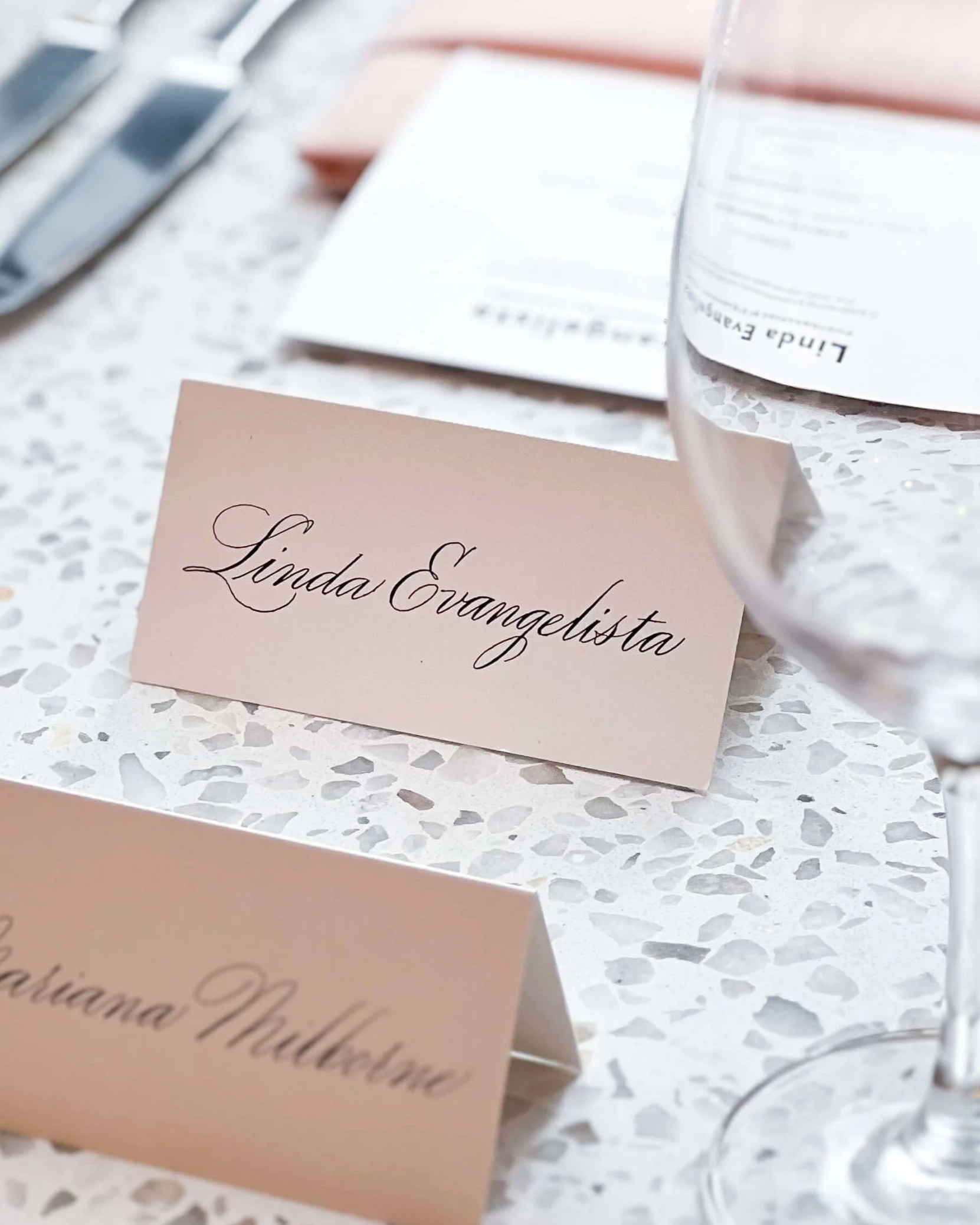 A white place card with the names written in calligraphy, placed on a white terrazzo table at a  luncheon event.