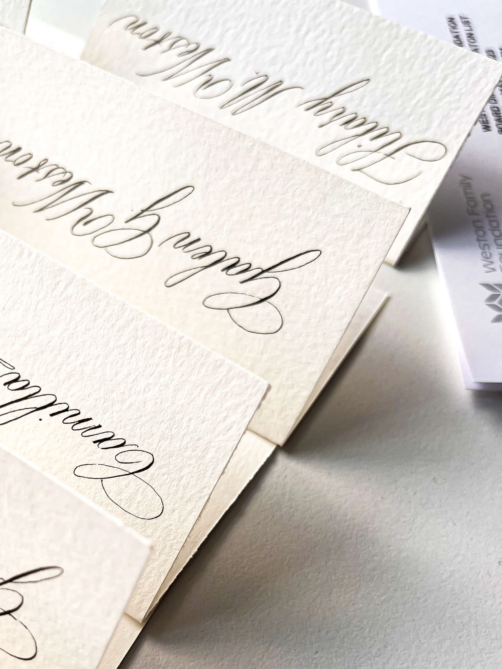 Multiple wedding invitations or place cards with elegant cursive writing on textured white paper, arranged in overlapping manner on a white surface.