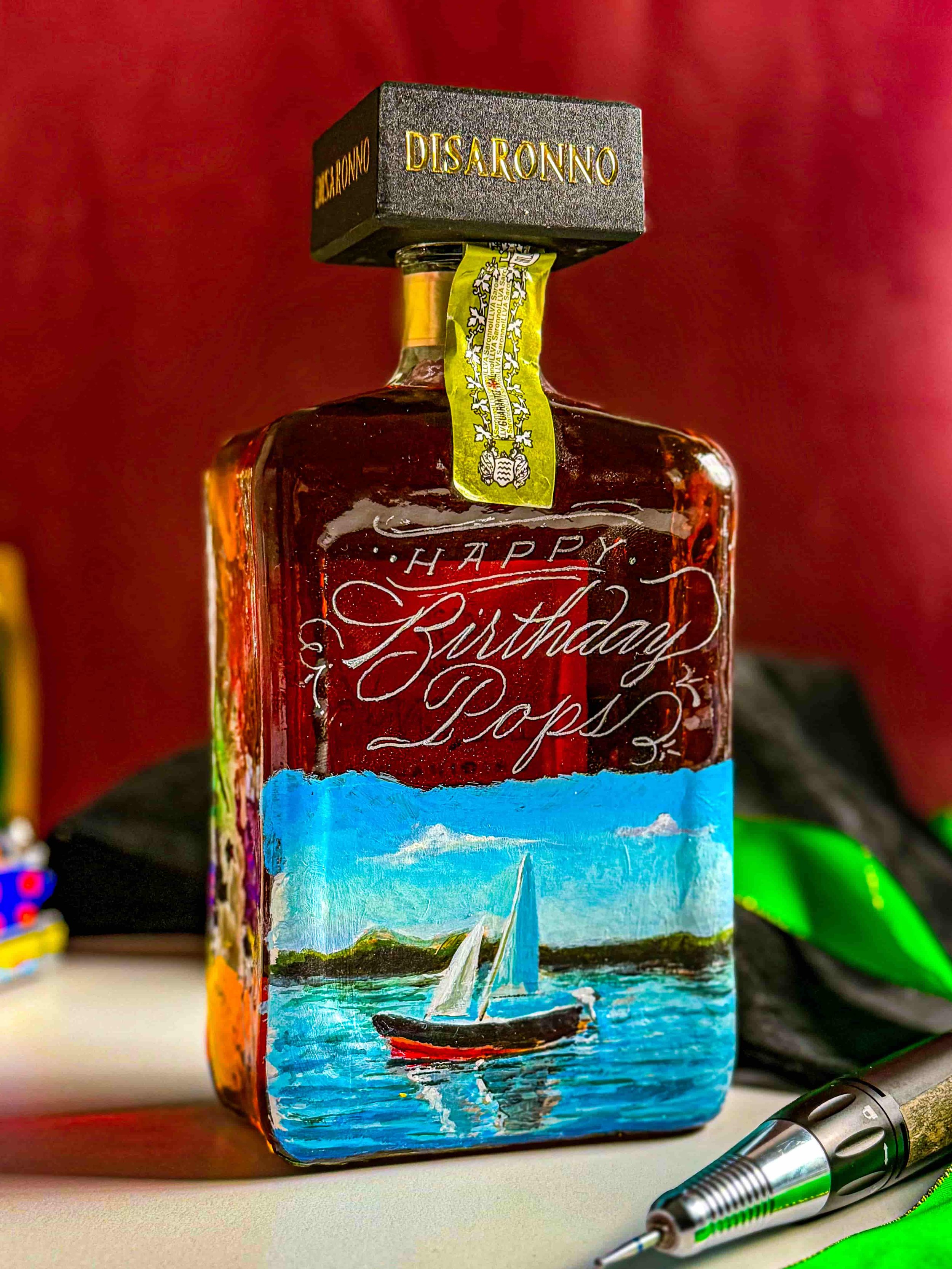 Decorated glass bottle with a boat painting and the hand engraved words 'Happy Birthday Pops,' topped with a black cube labeled 'Disaronno.'