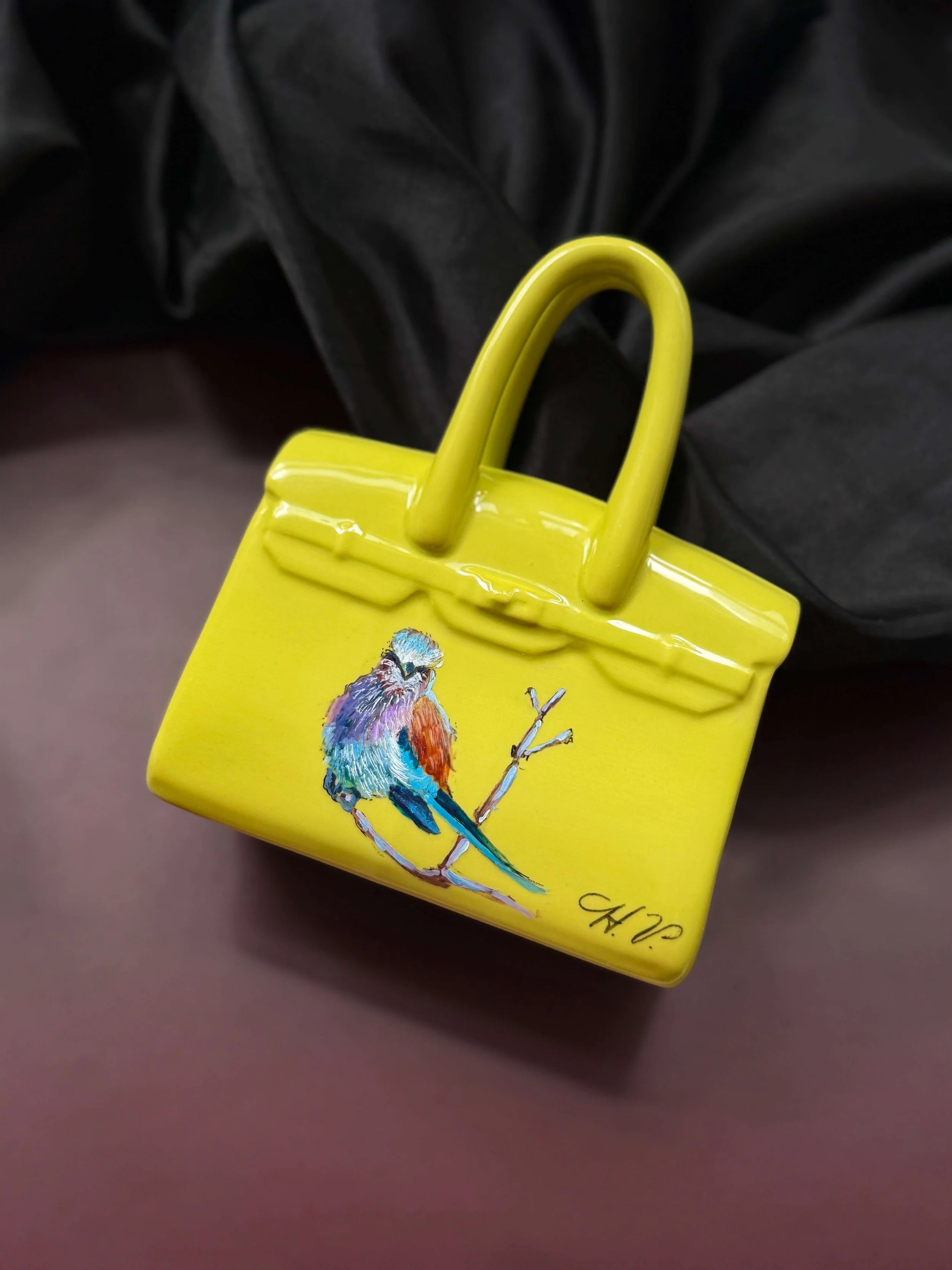 A small yellow purse with a painted bird on the front, perched on a branch, and a signature at the bottom.