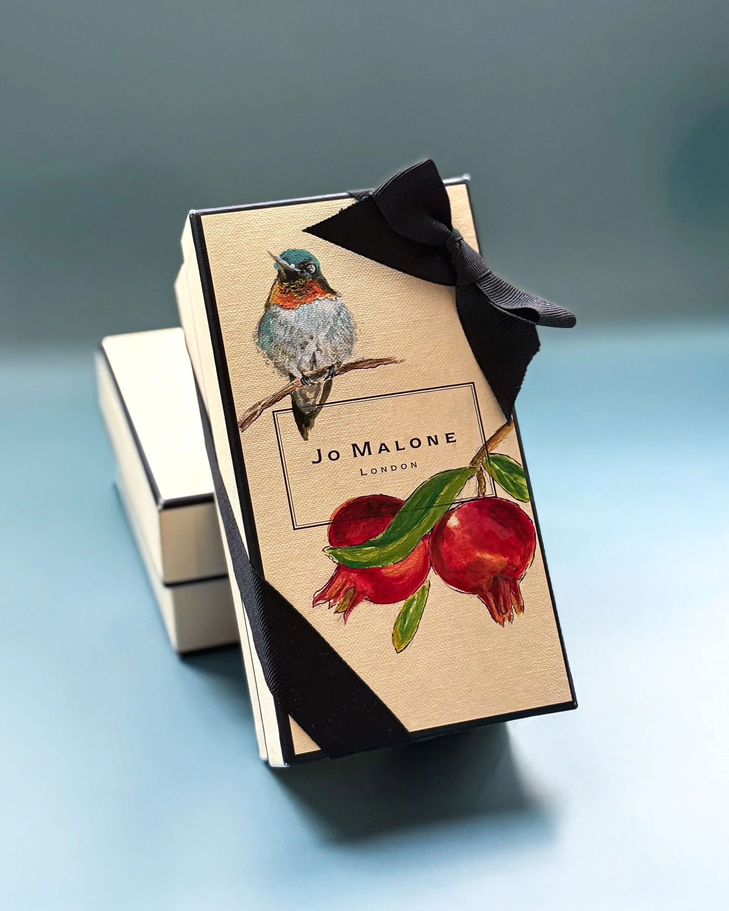 A decorative gift box with a Jo Malone London label, featuring an watercolor illustration of a bird perched on a branch with red pomegranate fruits, wrapped with a black ribbon.