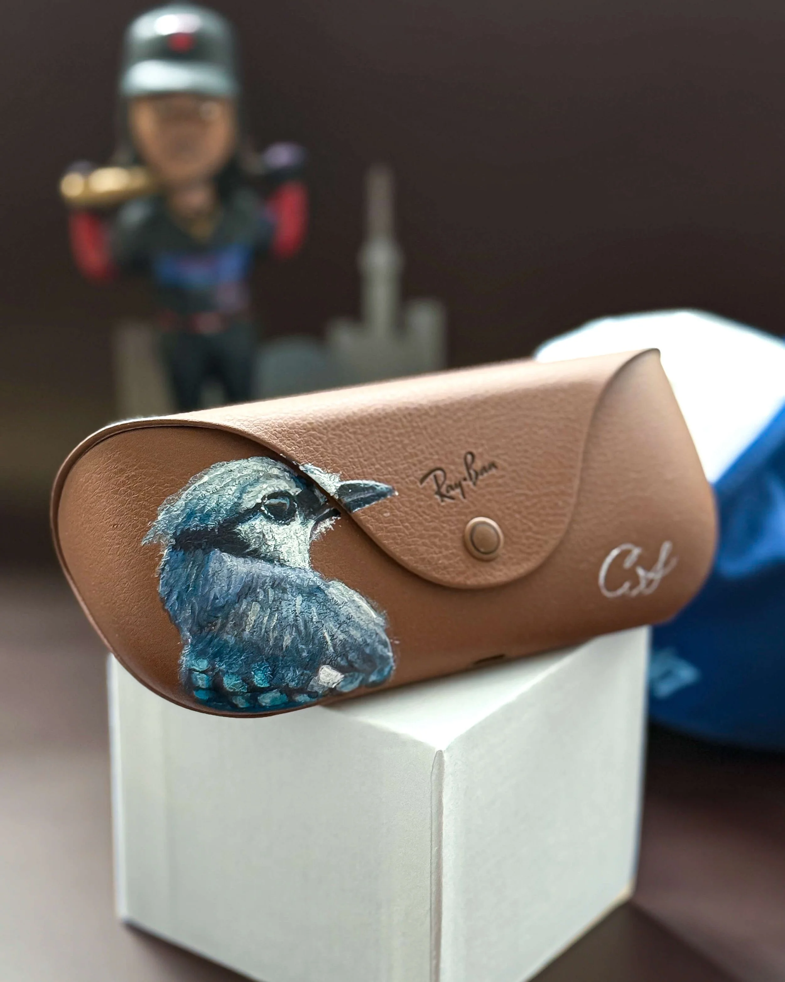 Close-up of a tan Ray-Ban eyeglasses case with a painted blue and gray bird, placed on a white box. In the background, a blurred figurine of a person in a uniform with a helmet and a cityscape silhouette.
