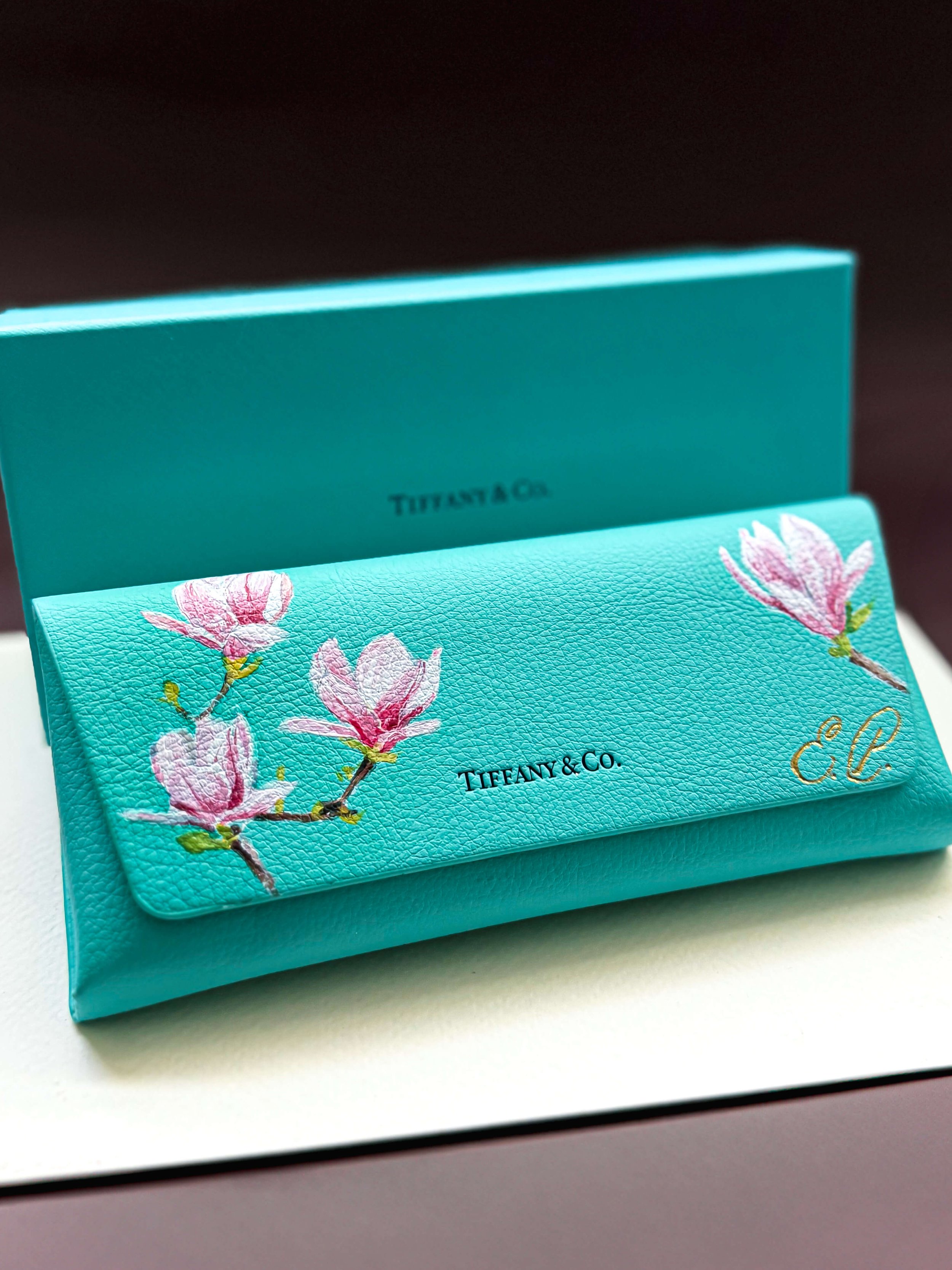 A turquoise leather eye glasses case with pink floral hand painting and gold  foiled initials, placed on a white surface with a Tiffany & Co. box in the background.