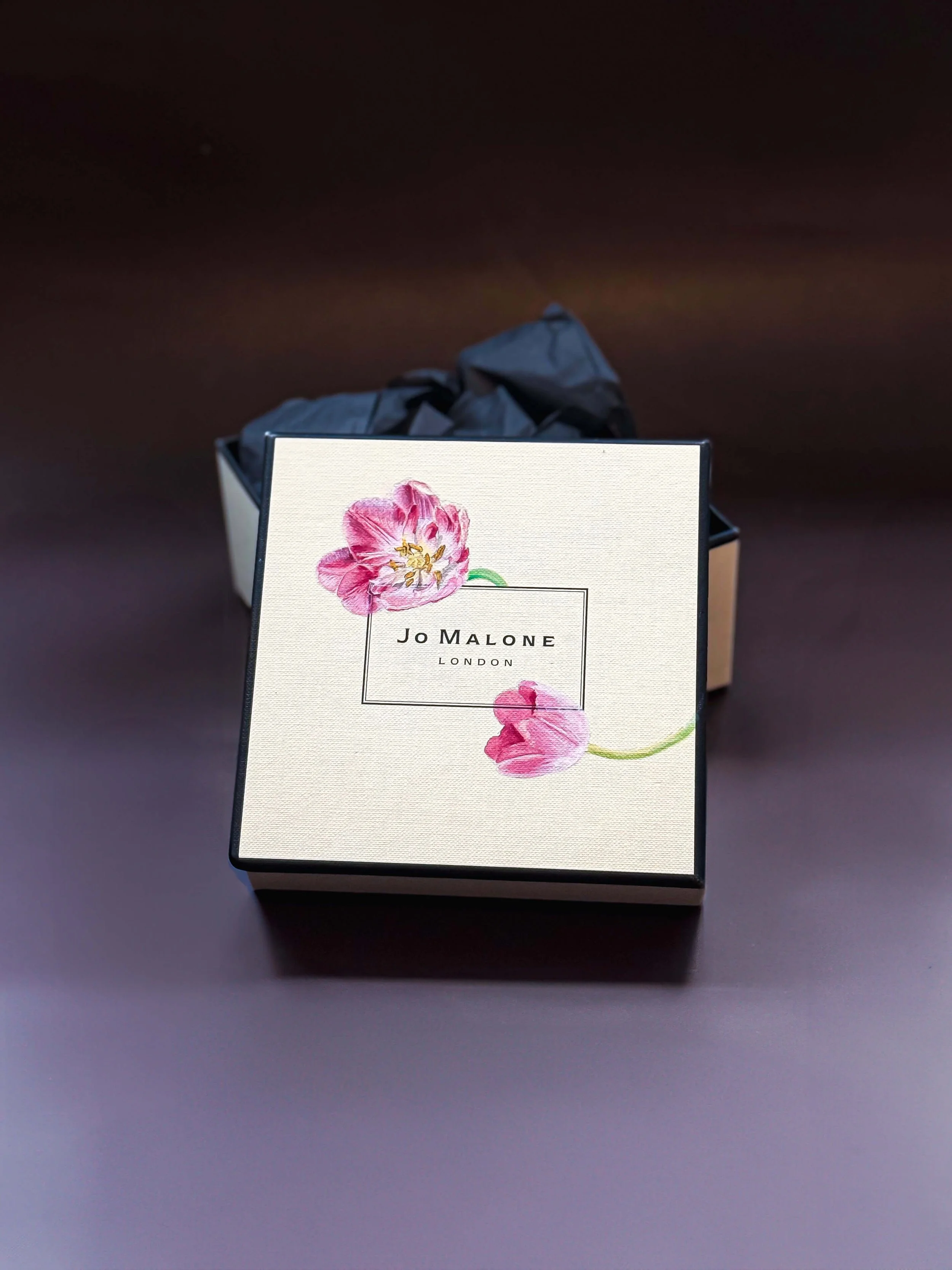 A Jo Malone London gift box with floral design, partially opened with black tissue paper inside, placed on a dark surface.
