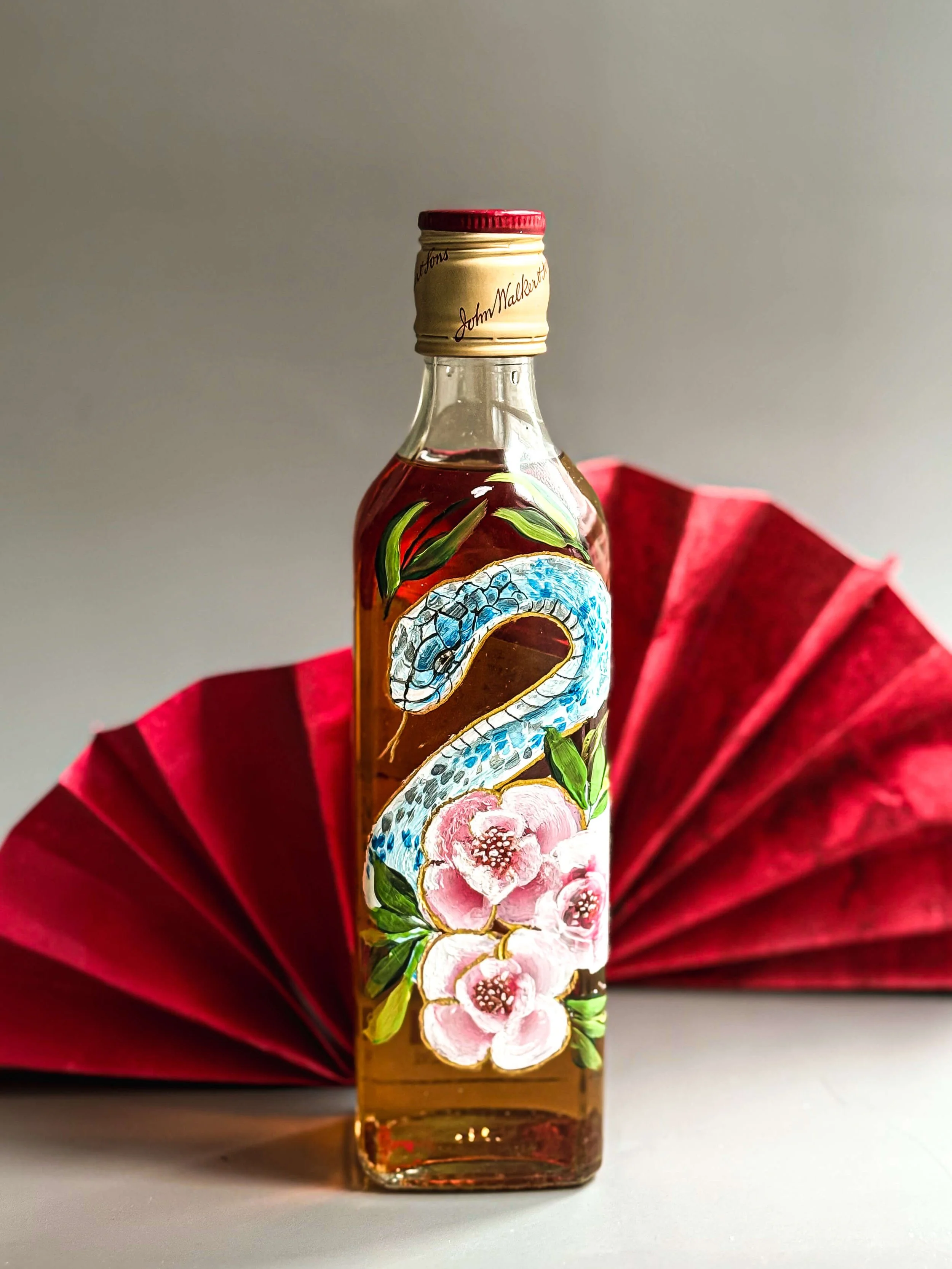 A small glass bottle with a tan and red cap, decorated with colorful paintings of a snake, pink flowers, and green leaves, placed in front of a folded red paper fan.