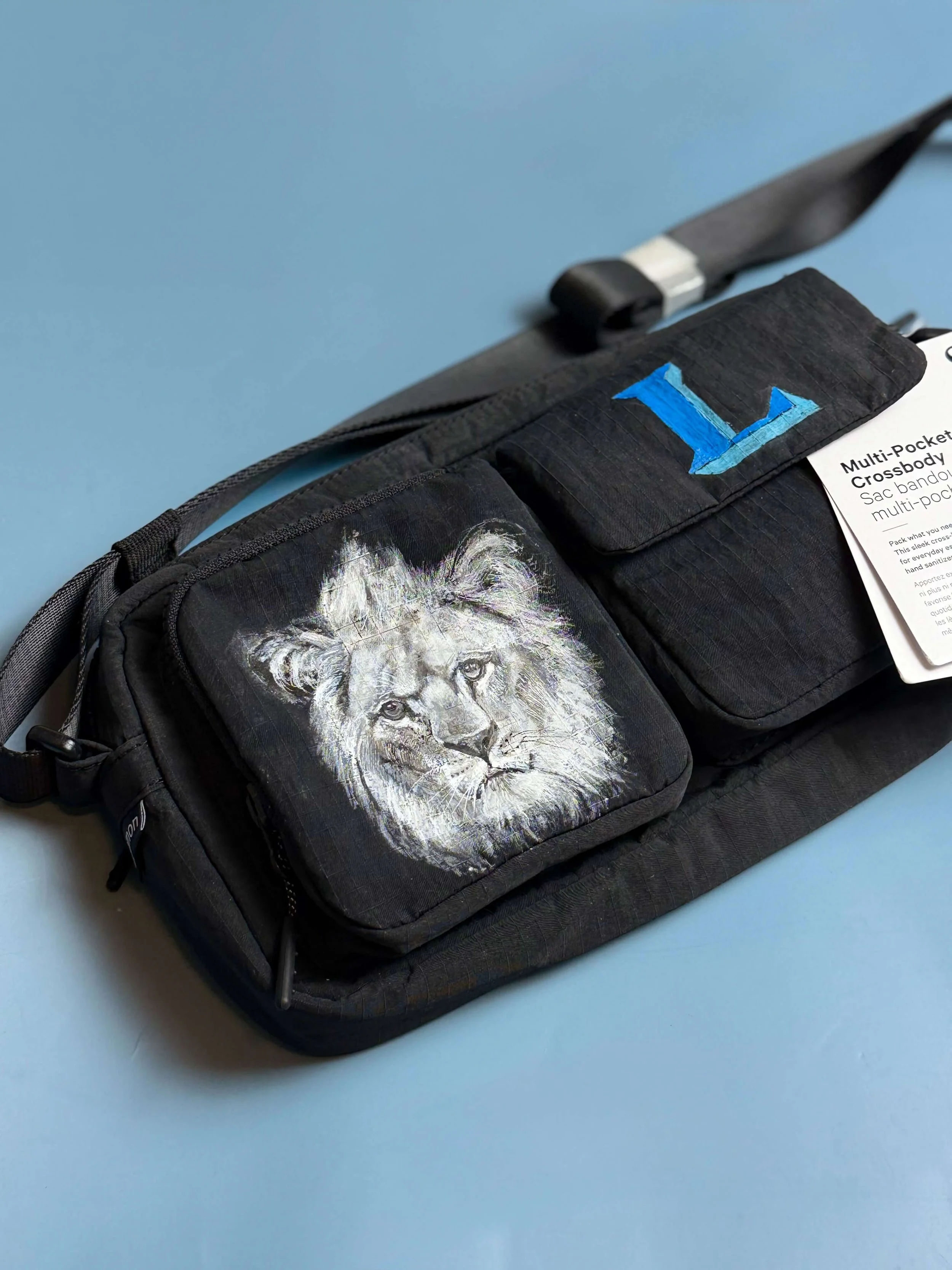 Black crossbody bag with a hand-painted lion's face on the front pouch and a hand painted monogram 'L' on the flap, placed on a light blue surface.