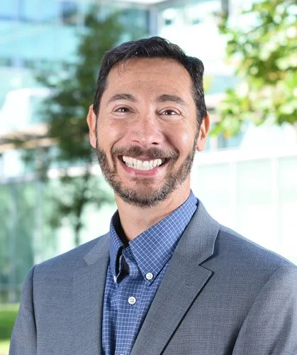 A man with dark hair and a beard, wearing a blue checkered shirt and a gray blazer, smiling outdoors with a blurred background of trees and buildings.
