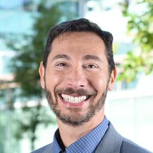 Headshot of a smiling man in business attire outside with greenery in the background.