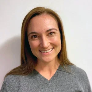 A woman with shoulder-length brown hair and a gray top, smiling at the camera, standing against a plain white background.