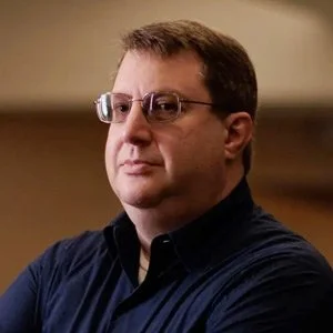 A man with short brown hair, glasses, and a serious expression wearing a dark shirt.