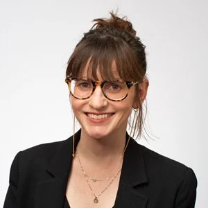 A woman with brown hair in a bun, wearing glasses, a black blazer, and layered necklaces, smiling at the camera.