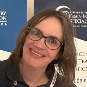 Smiling woman with glasses and shoulder-length hair at a professional event with banners in the background.