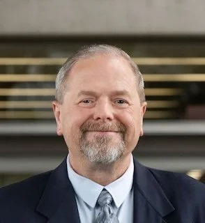 Portrait of a man in a blue suit, white shirt, and light tie, smiling against a medical building.
