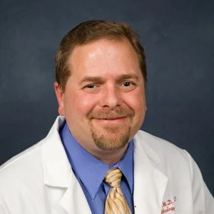 Portrait of a man in a white coat, light blue shirt, and beige tie, smiling against a dark background.