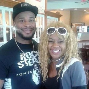 A man and woman posing together indoors, both smiling. The man is wearing a black cap and a black T-shirt, and the woman has blonde curly hair, white sunglasses on her head, and a gray jacket.