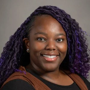 A young woman with dark skin and curly purple-tinted hair smiling at the camera.