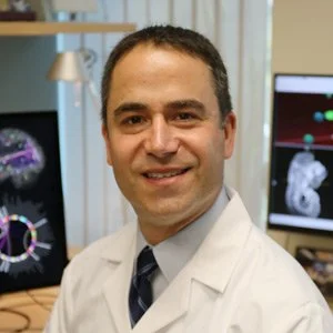 A man in a white lab coat smiling in a medical or research setting with computer screens displaying medical imaging and brain scans in the background.