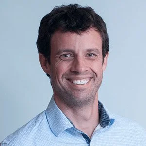 A smiling man with dark, curly hair wearing a light blue button-up shirt against a plain light blue background.