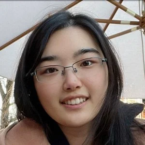 A woman with black hair and glasses smiling at the camera, sitting outdoors under a large umbrella.