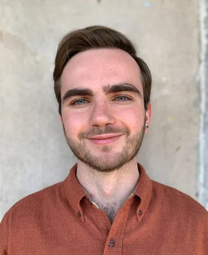 Young man with brown hair, beard, and blue eyes smiling in front of a plain wall, wearing a brown button-up shirt.