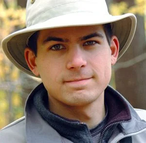 Close-up of a man wearing a beige hat and outdoor clothing, standing outdoors with trees in the background.