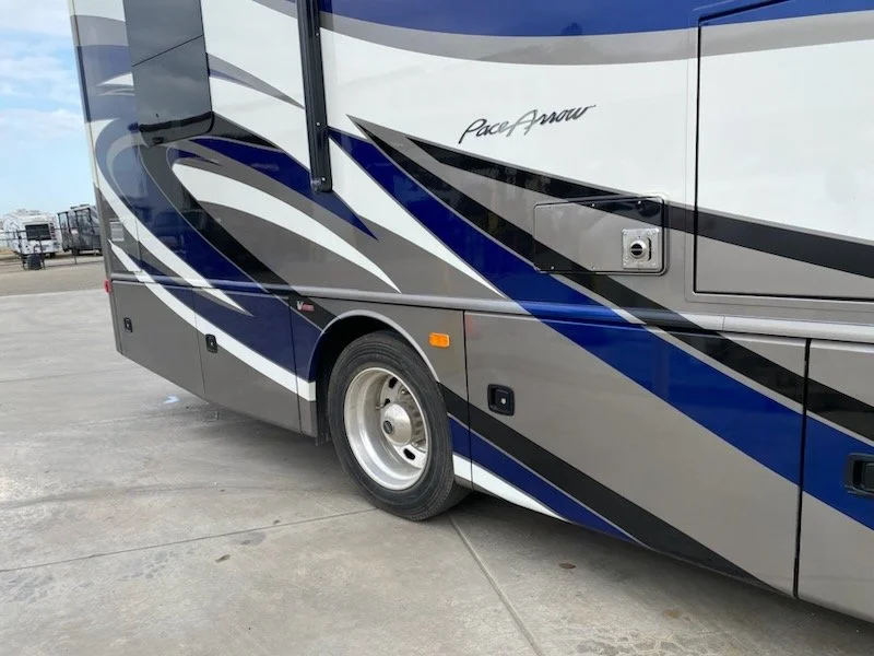 Close-up of the side of a recreational vehicle (RV) with a blue, black, and gray design, and the words "Pace Arrow" written on it, parked on a concrete lot with other RVs in the background.