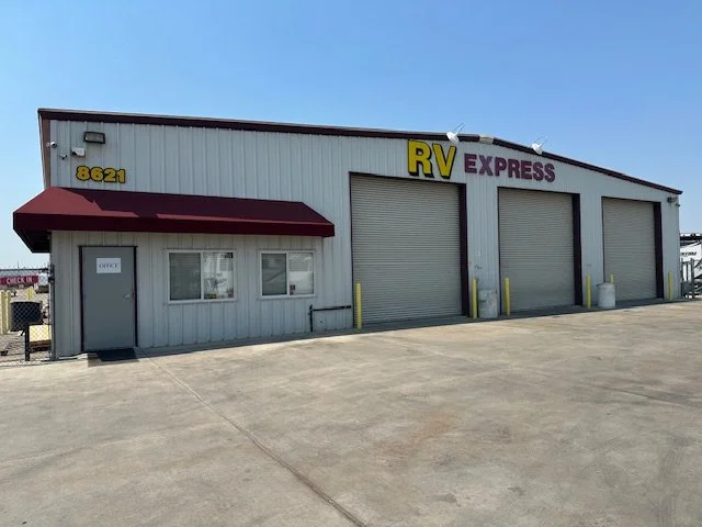 An industrial building with a sign that reads RV Express, three large garage doors, a small door with a red awning, and a concrete parking lot in front.
