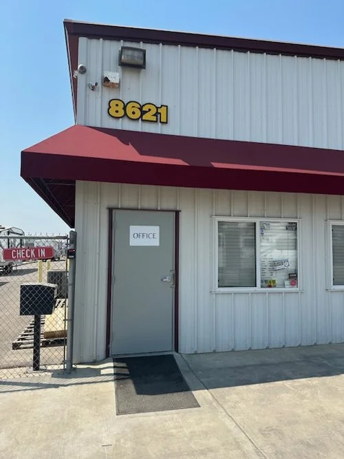 Exterior of a small building with the number 8621 above and an office door with a sign that says 'Office.' There is a window, a black doormat, and a chain-link fence with a sign that says 'Check In.'