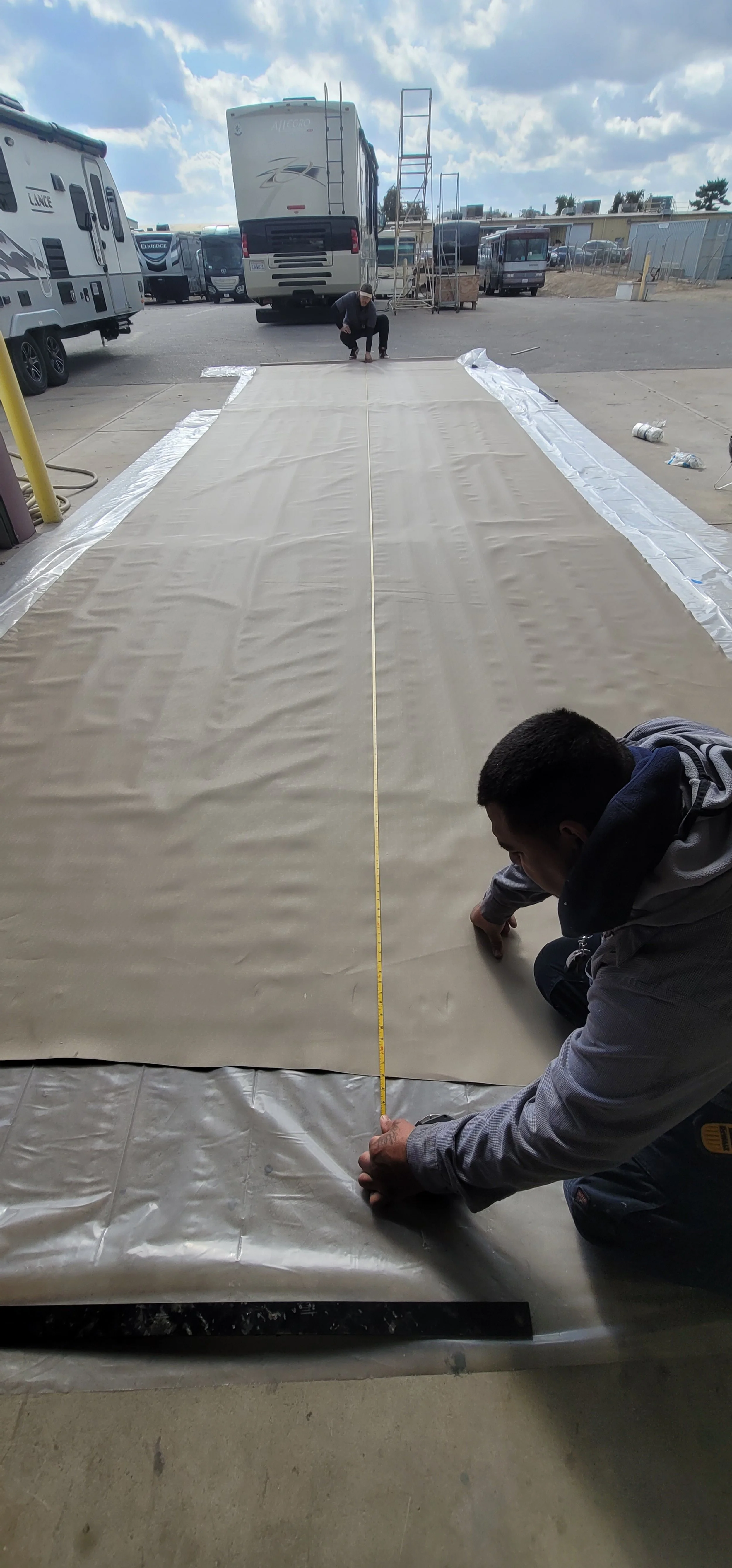 Two workers measure and prepare a large sheet or tarp on the ground outside, with multiple RVs and trucks parked in the background under cloudy skies.