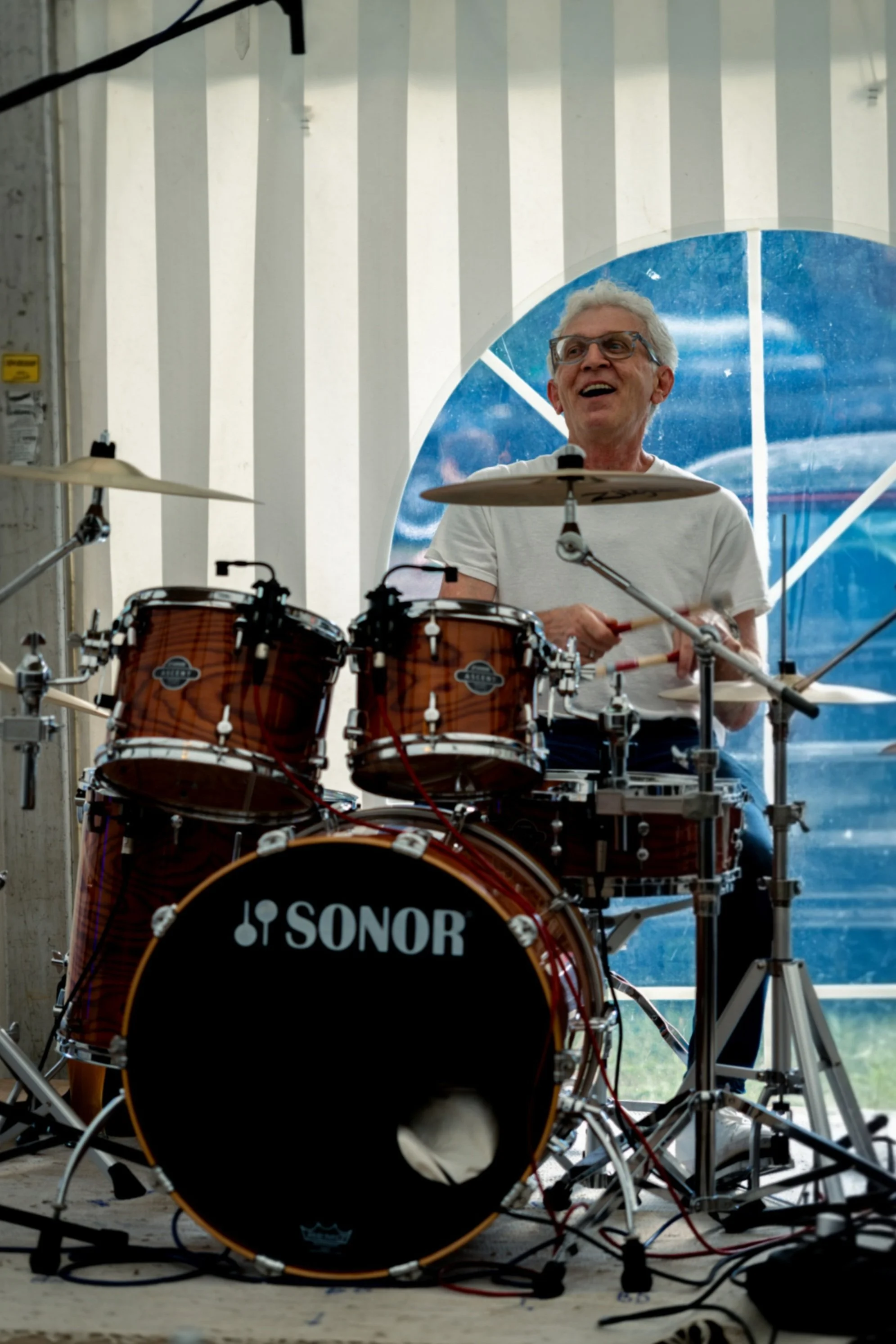 Ein Mann mit Brille sitzt an einem Schlagzeug der Marke Sonor. Er trägt ein weißes T-Shirt und lacht. Im Hintergrund sieht man die Rückwand eines Festzeltes.