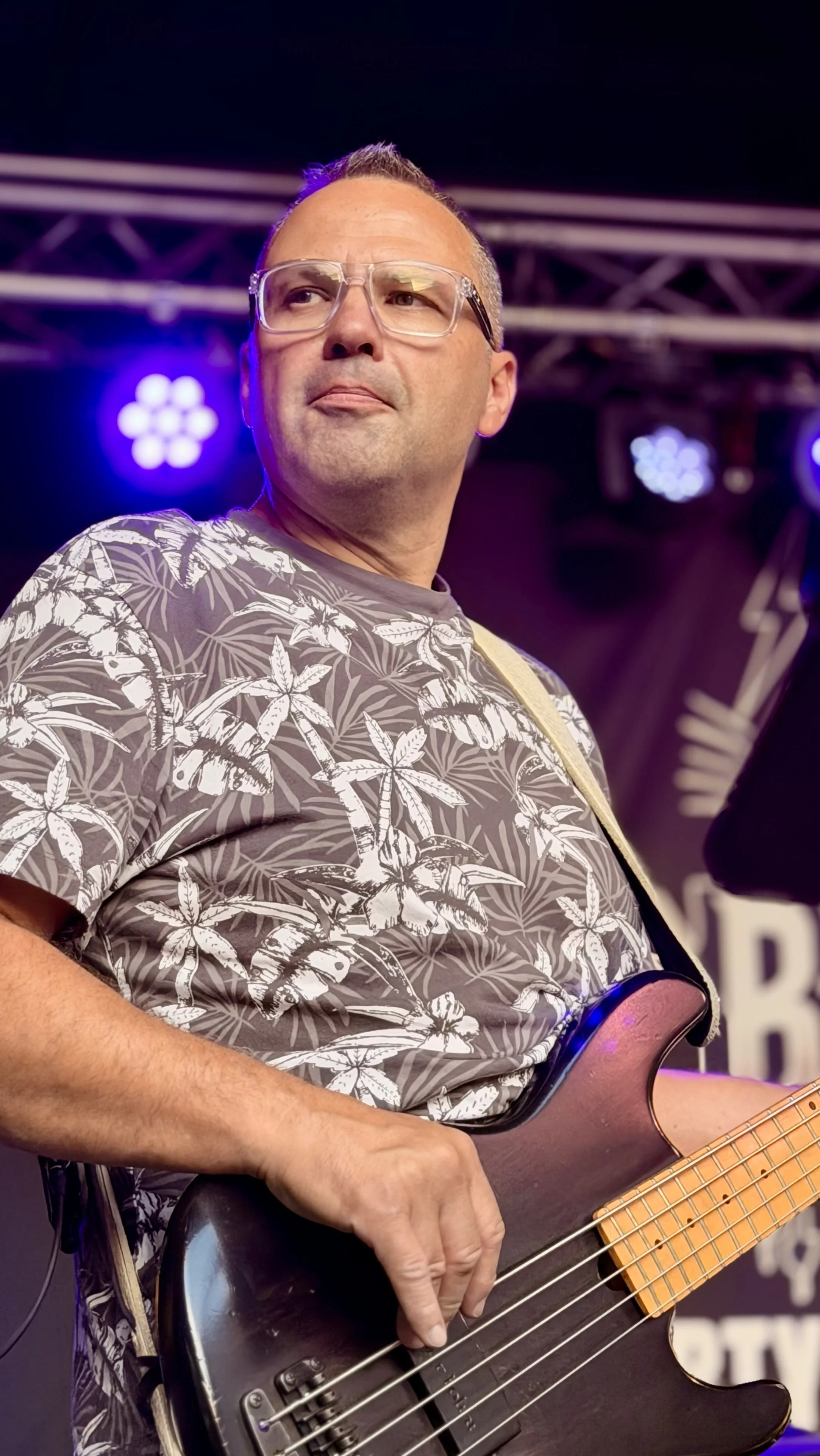 Mann spielt E-Bass auf einer Bühne mit blauen und weißen Lichtern, trägt ein T-Shirt mit Palmenmuster, trägt Brille und hat kurzes, braunes Haar.