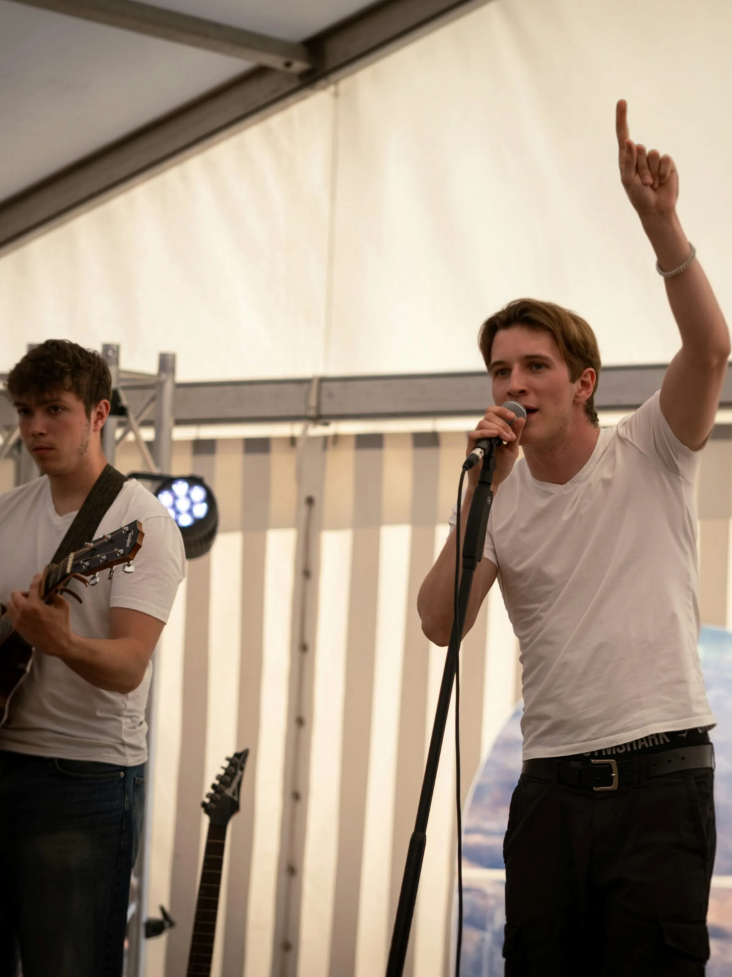 Zwei junge Männer beim Auftritt auf einer Bühne in einem Festzelt. Einer singt in ein Mikrofon, der andere spielt E-Gitarre.