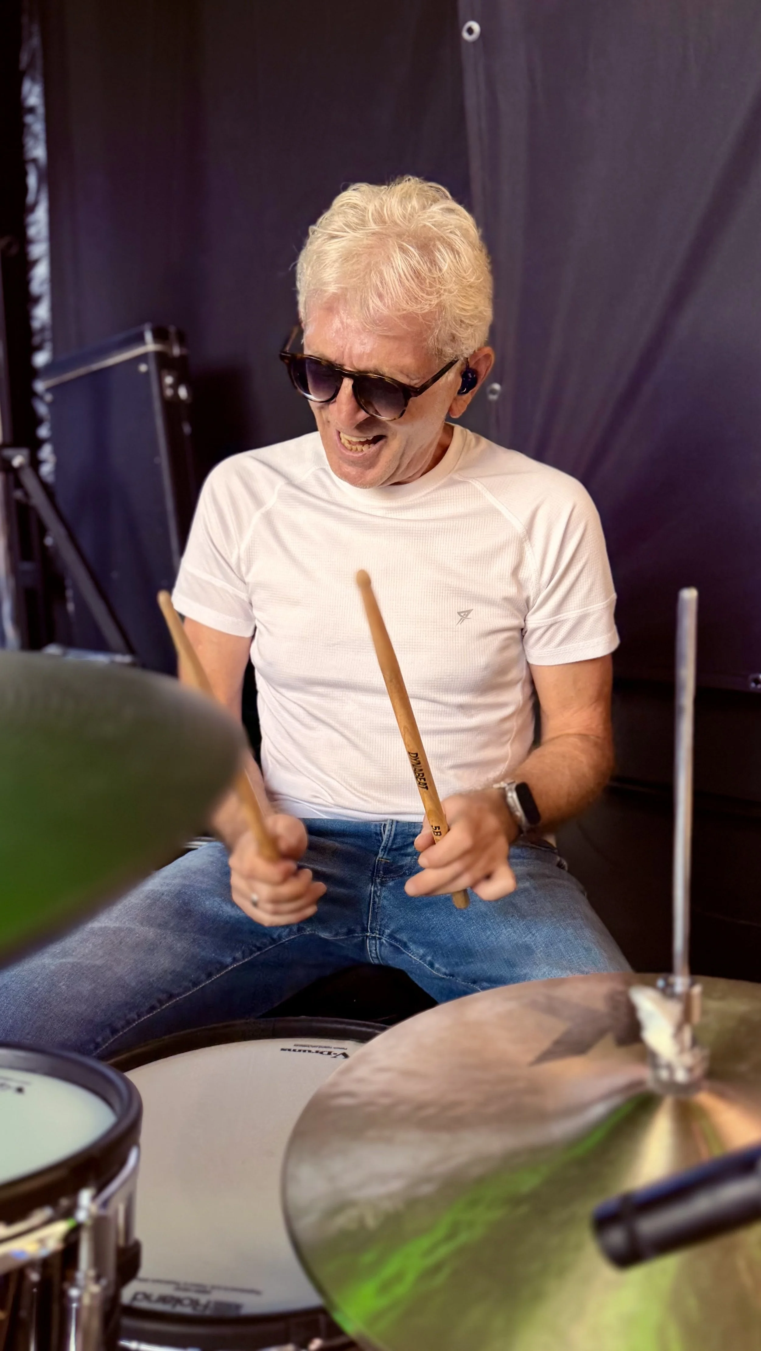 Ein Mann trägt Sonnenbrille, spielt auf einem Drumset, lächelt und trägt ein weißes T-Shirt. Im Hintergrund eine dunkle Wand.