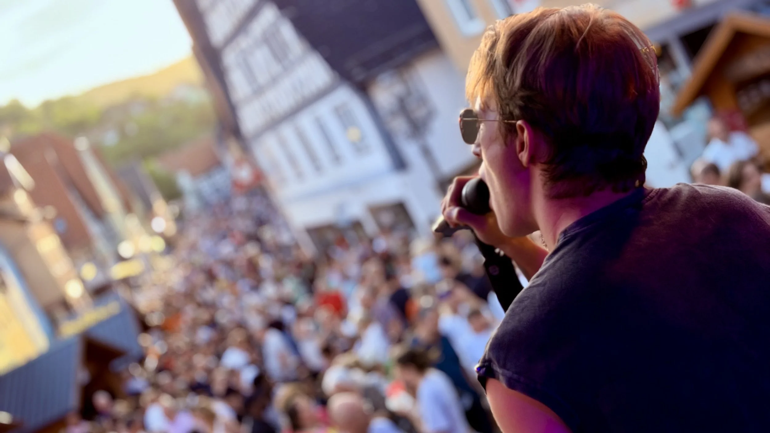 Ein junger Mann mit dunkelblondem Haar trägt Sonnenbrille und hält ein Mikrofon bei einem Festival im Freien, umgeben von einem großen Publikum.