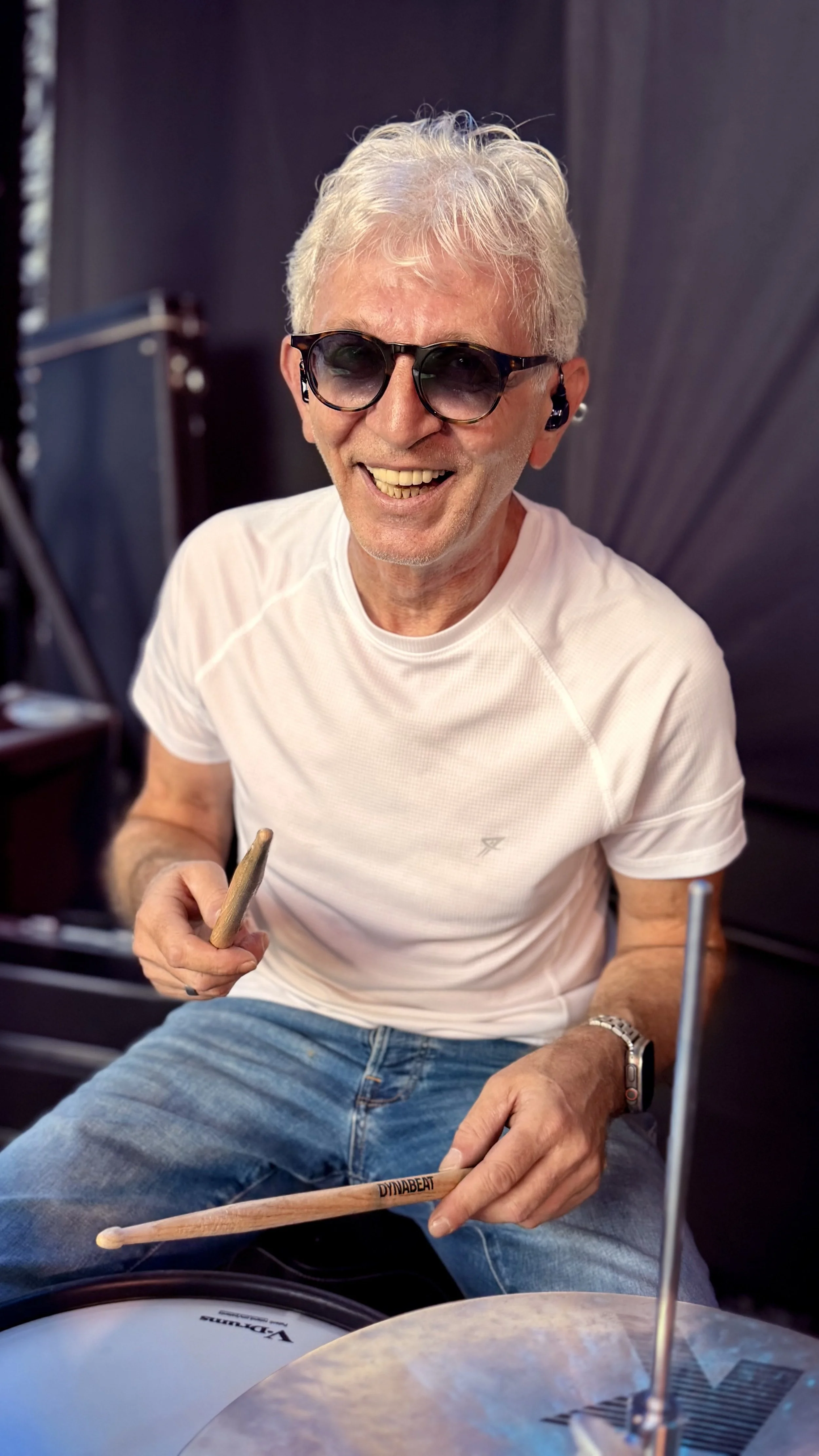 Ein grinsender Mann mit grauem Haar, dunklen Sonnenbrille und weißen T-Shirt beim Musizieren, er hält ein Drumstick in der Hand, sitzt vor einem Schlagzeug, im Hintergrund dunkle Vorhänge.