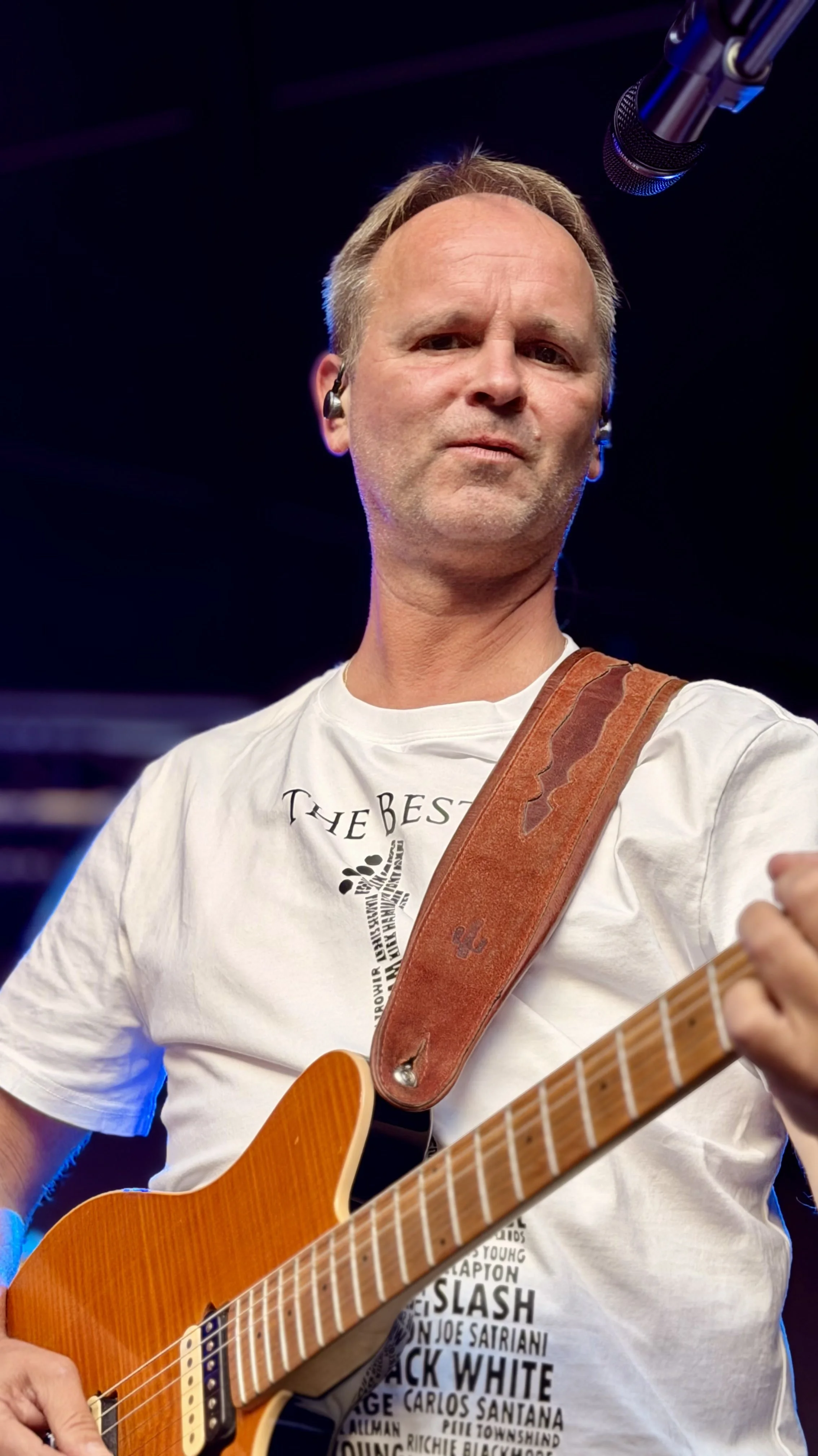 Ein Mann spielt Gitarre auf einer Bühne, trägt ein weißes T-Shirt hat einen braunen Gitarrengurt.
