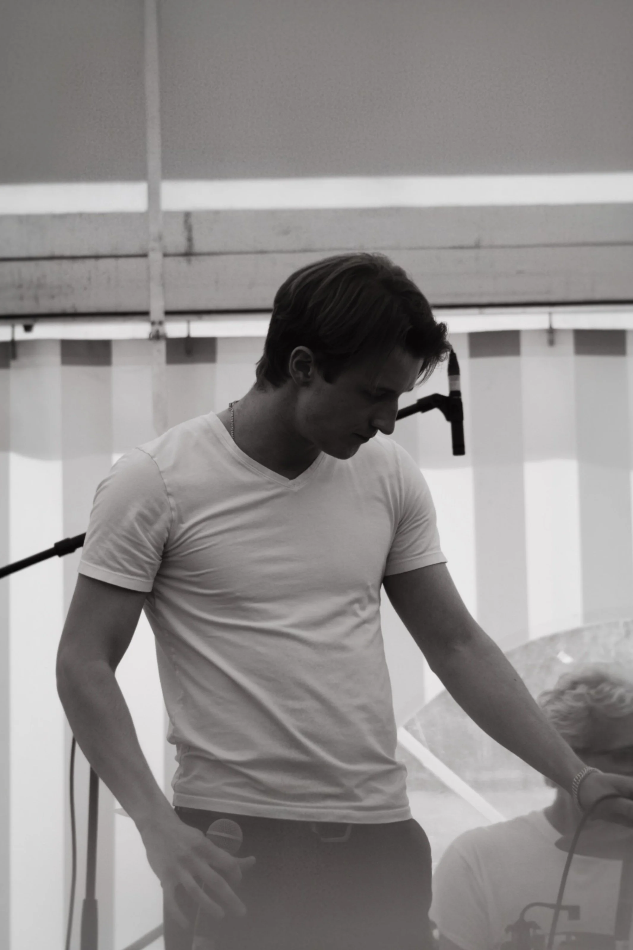 Schwarz-weiß-Foto eines jungen Sängers in weißem T-Shirt, der nach unten schaut, im Hintergrund die Rückwand eines Festzeltes.