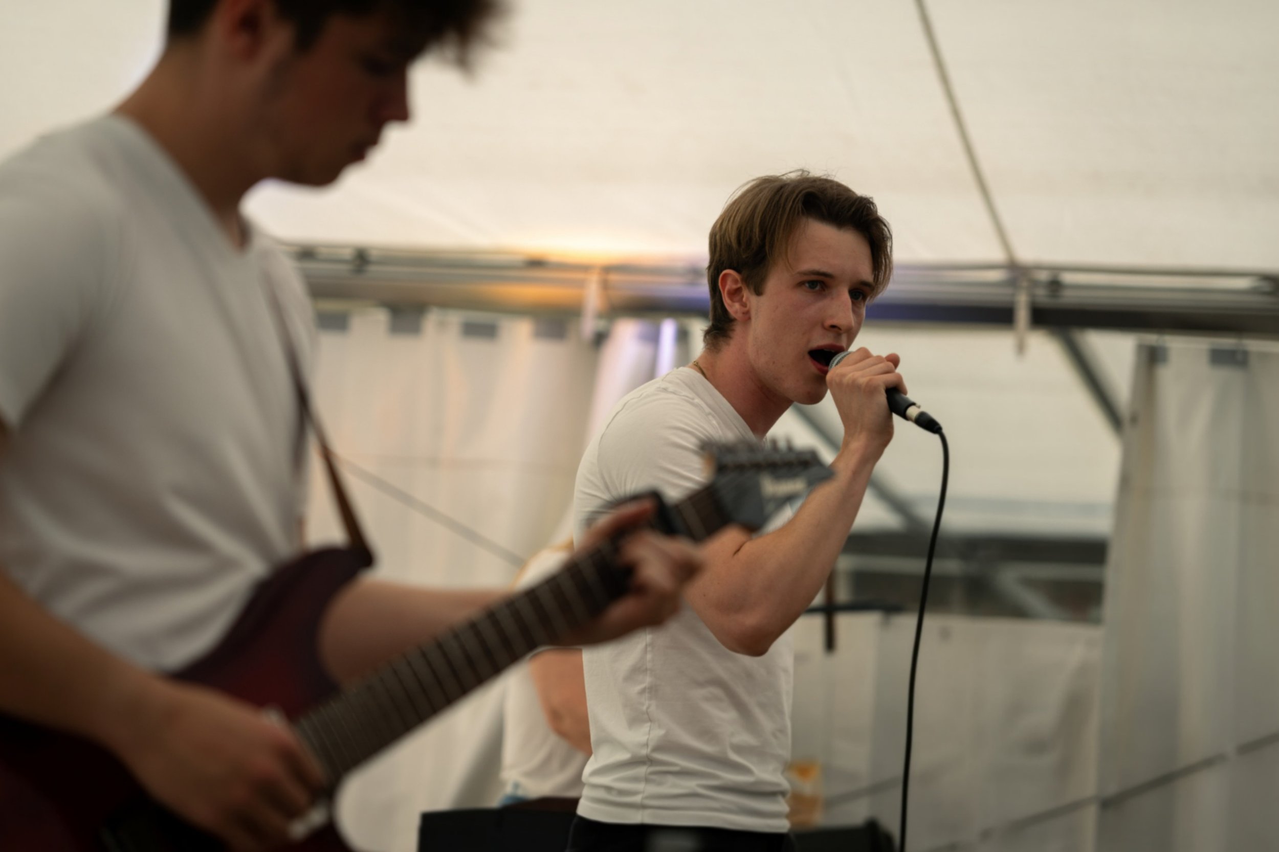 Zwei junge Männer spielen Musik in einem Festzelt, der eine singt mit einem Mikrofon, der andere spielt Gitarre.