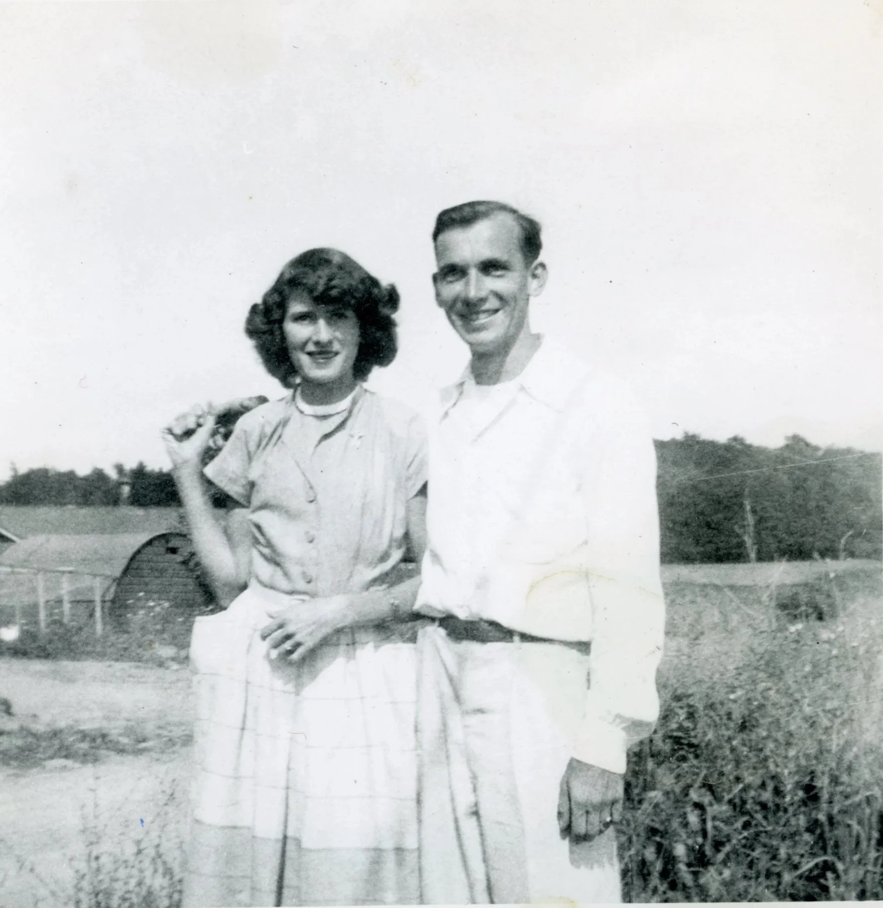 A black and white photo of a smiling man and woman standing outdoors in a rural setting, with fields and trees in the background.