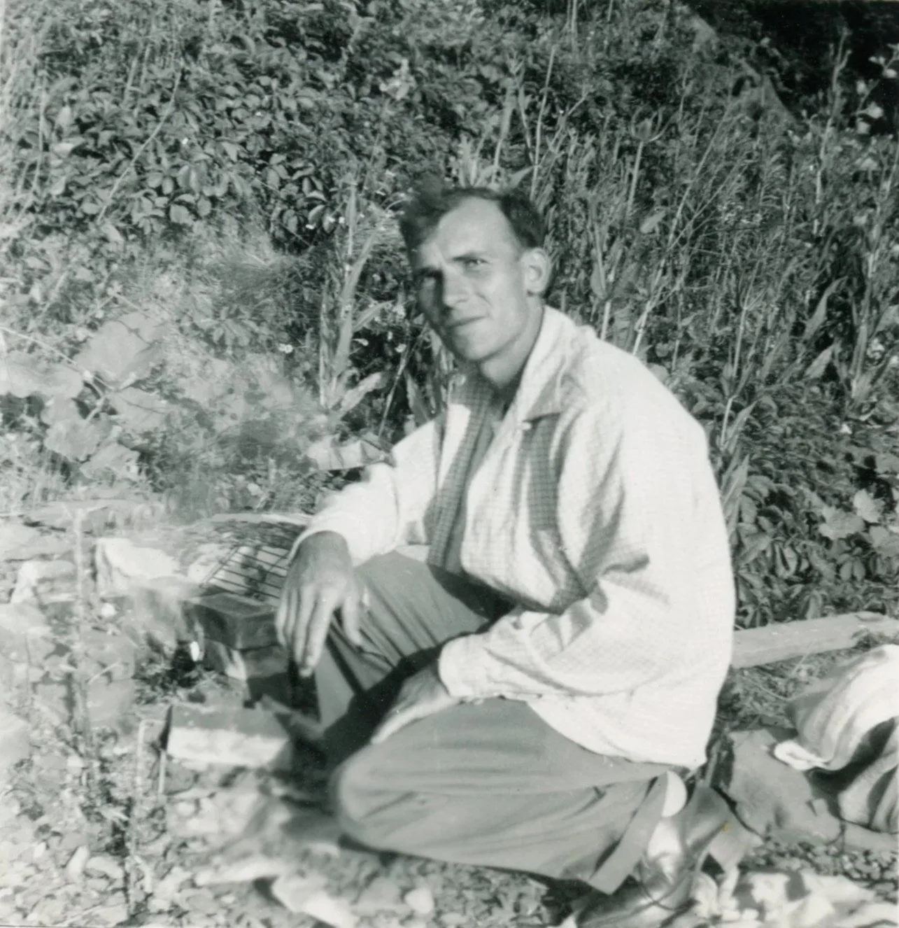 A man kneeling on the ground in a field of tall plants, possibly a farm or garden, holding a wooden object and looking at the camera.