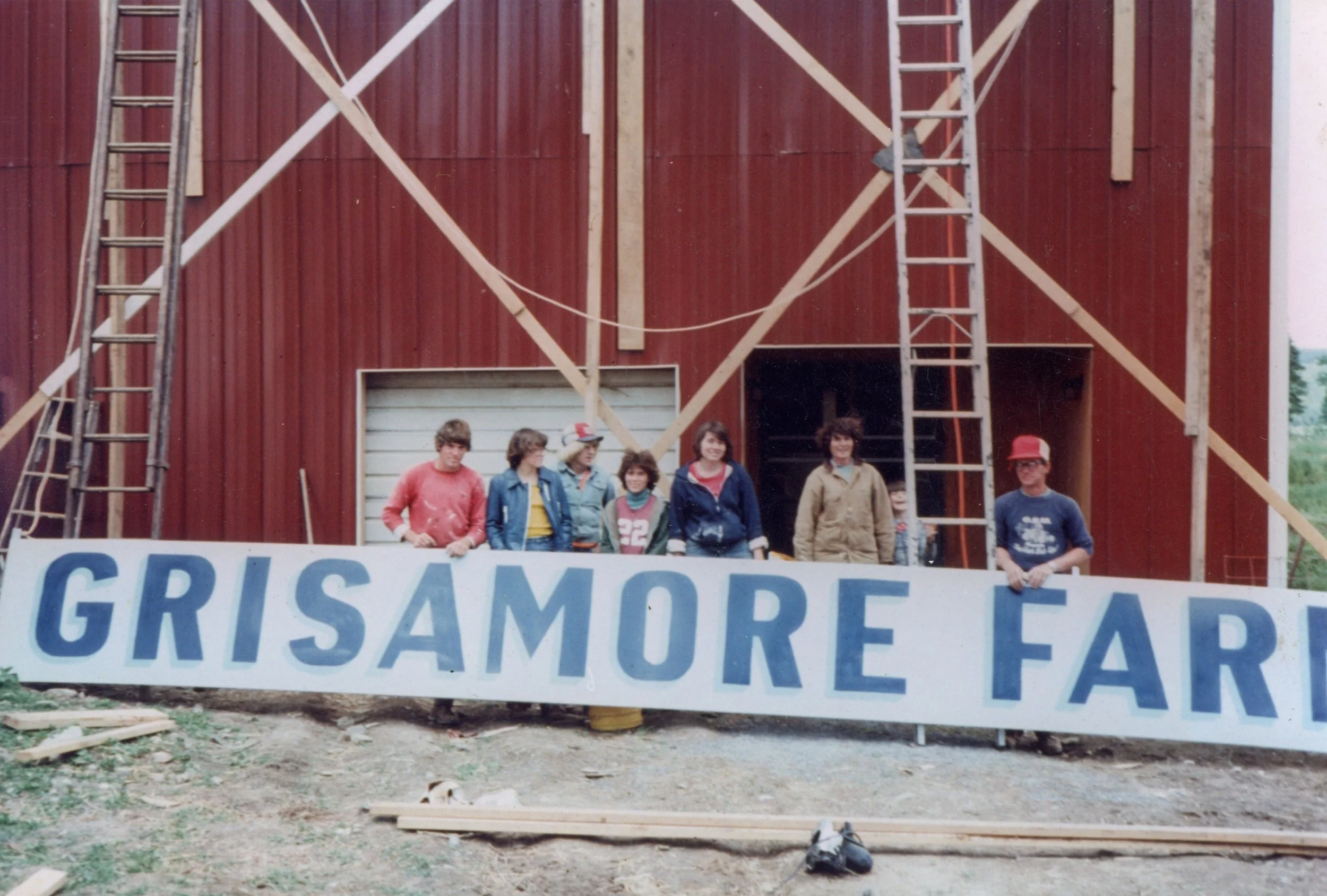 Group of children and teenagers standing behind a large sign that reads 'GRISAMORE FARM' in front of a building under construction with ladders and wooden beams.