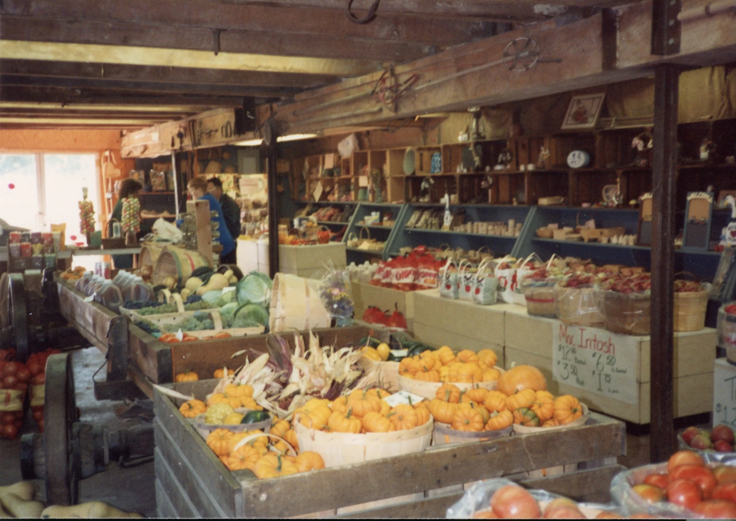 Inside a rustic farmers market with fresh produce such as pumpkins, onions, and vegetables on display. Shelves stocked with goods and two people shopping near the counter.
