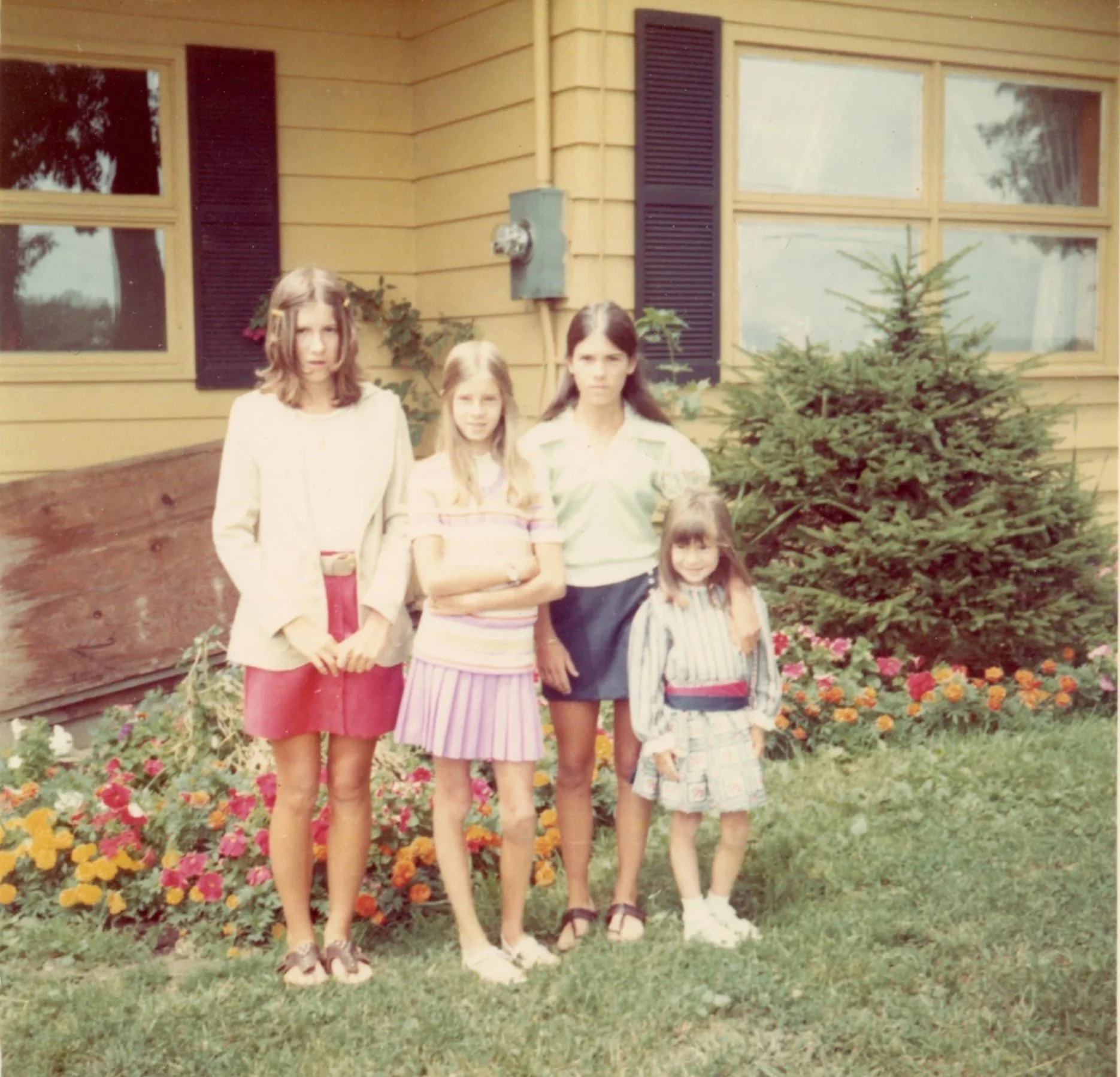 Four girls standing in front of a yellow house with a garden and flower bed, posing for a photo.