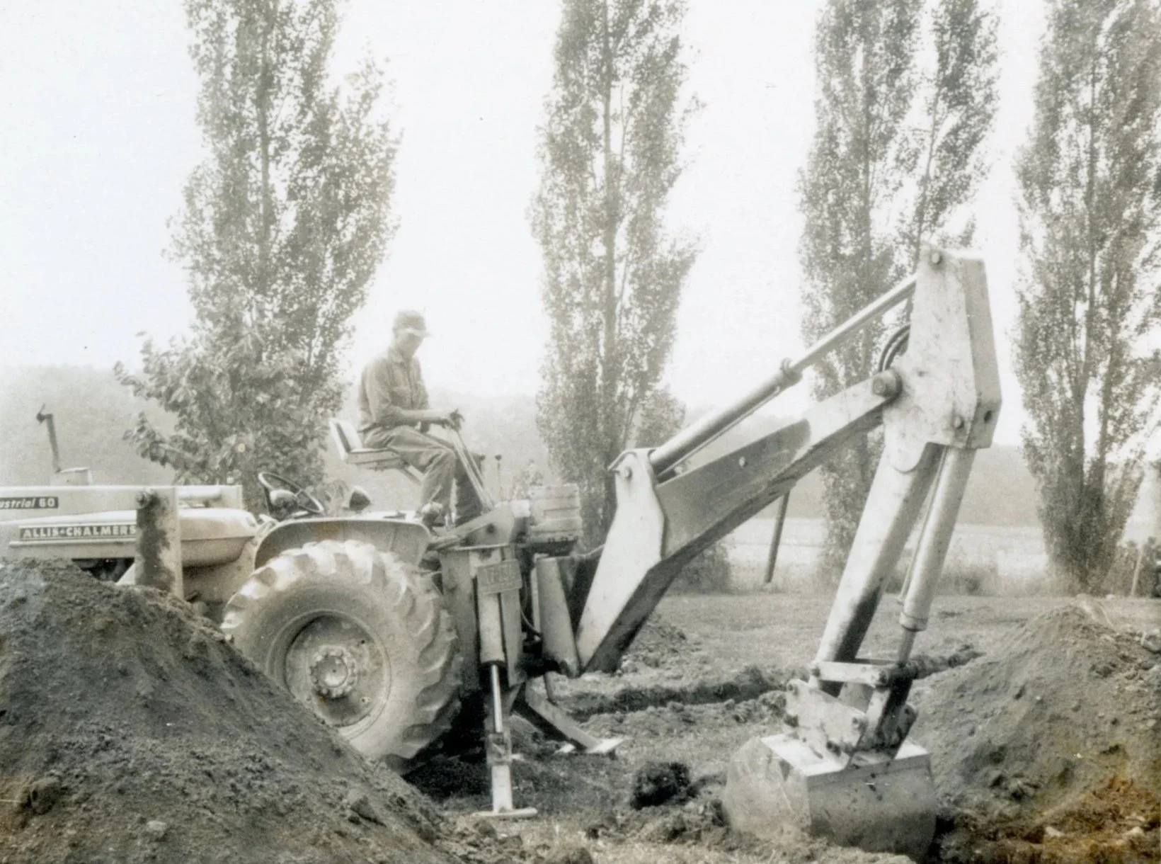 A black and white photo of a man operating a tractor with a front loader, digging into the ground outdoors. There are tall trees in the background.