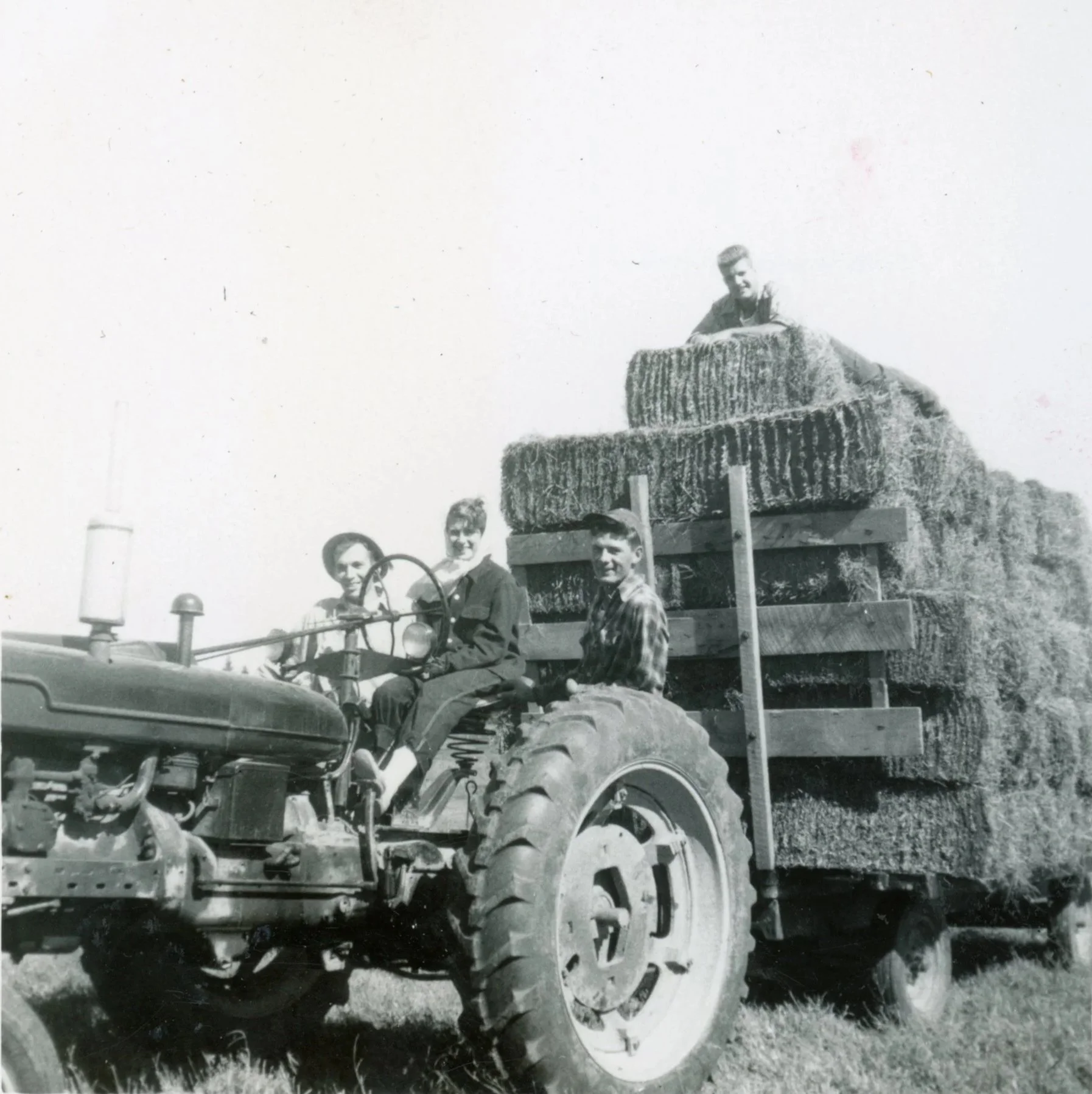 Black and white photo of four people on a tractor carrying a large load of hay bales, with three seated on the tractor and one on top of the hay.